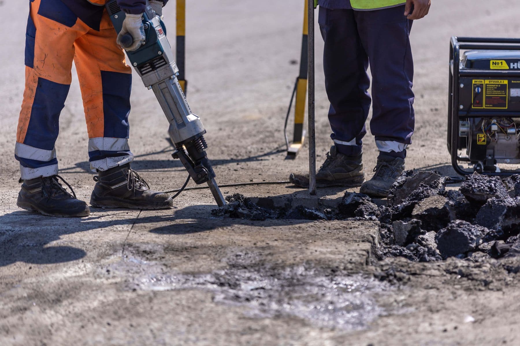 Two workers' legs beside a jackhammer breaking up a paved surface, with a small generator nearby.