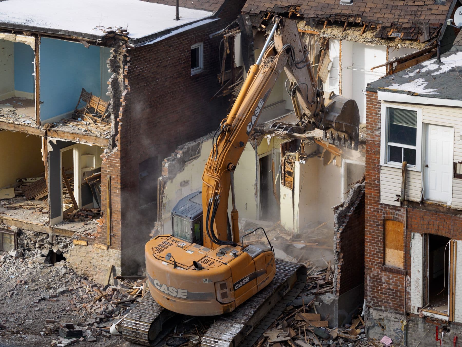 An excavator tearing down a row house between two others, with the inside rooms open to view.
