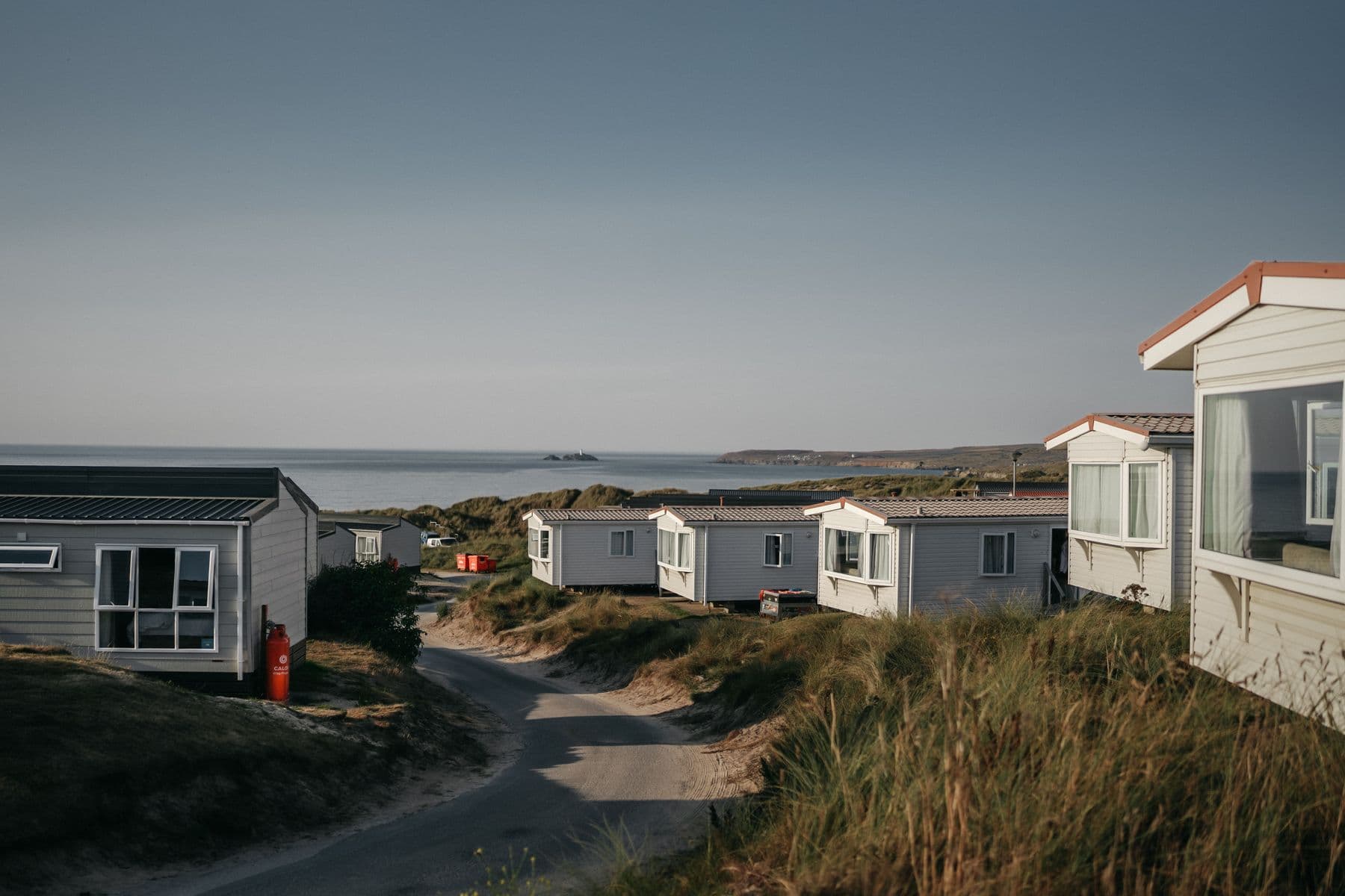 A row of white static caravans on a sandy track among dune grass, with the sea behind them.